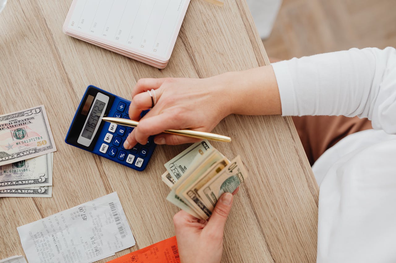 Person using a calculator and counting cash on a wooden table with receipts.